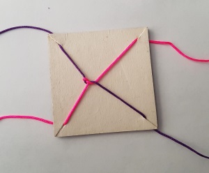 Photo of a square piece of card with a small slit diagonally at each corner. Each slit holds in place one end of a piece of string. There is a tangle of string in the middle of the square.