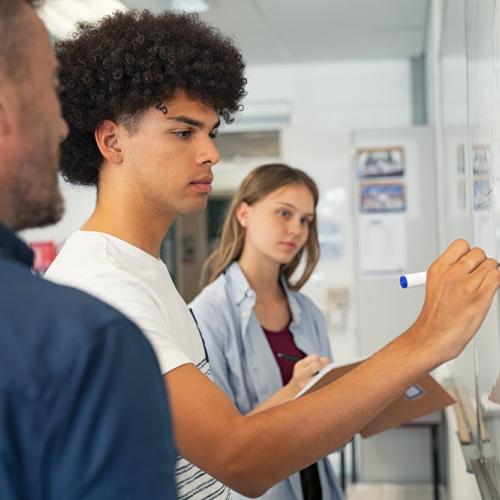 Several university age students standing at a whiteboard, with one guy writing on it