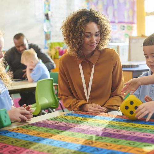 Teacher wearing brown at a table with primary school children, with a colourful grid on the table