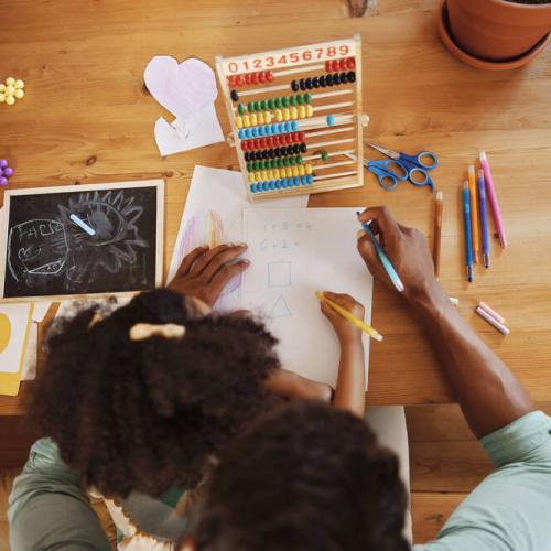 Overhead photo of girl and adult doing maths on a wooden desk