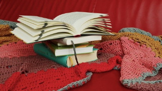 an open book on top of a pile of books, resting on a crochet blanket