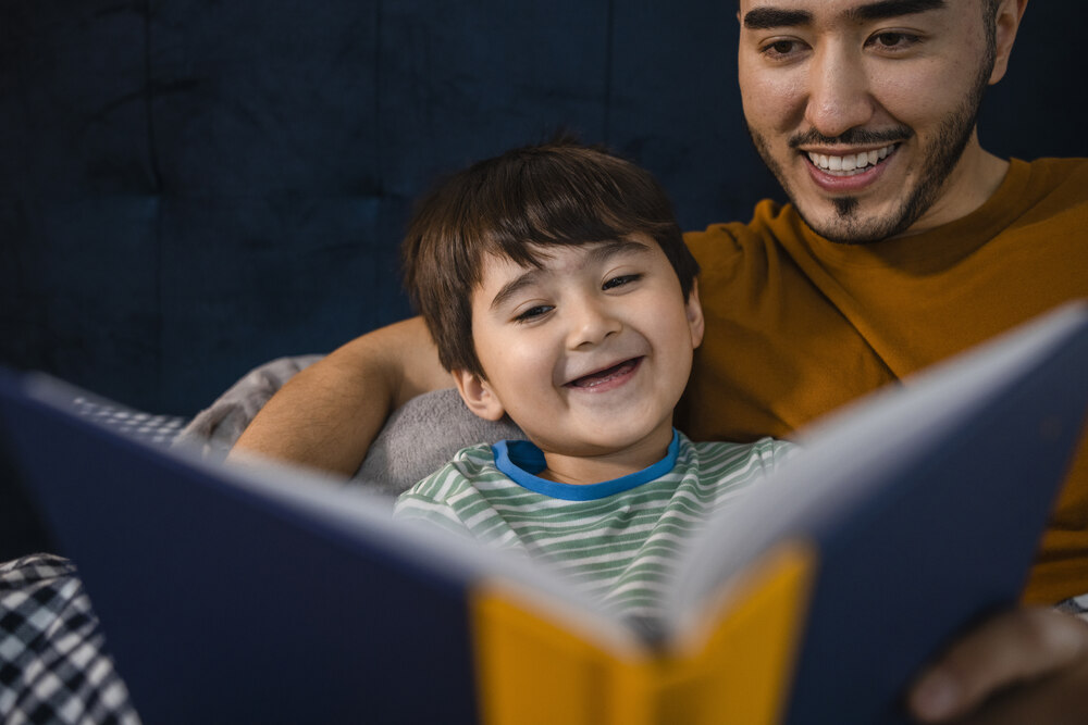 Small boy with a big smile silling in the lap of an older man, with a big book open in front of them