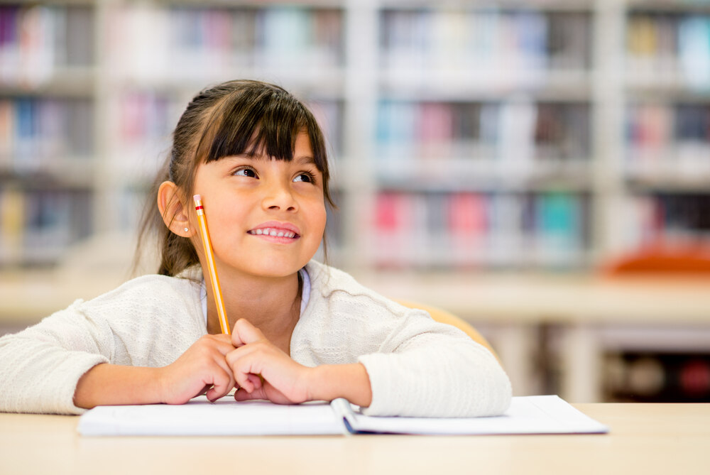 Young girl smiling while looking up and to her left, holding a pencil and sitting at a table with a notebook