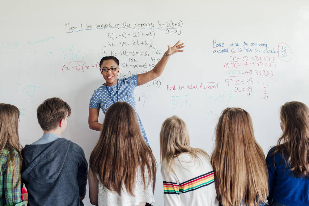 Classroom of secondary school students, with a teacher at the front of the class in front of a whiteboard with lots of colourful mathematics