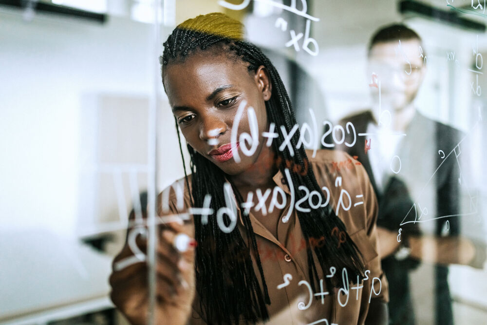 Photo looking through glass, of a woman writing maths in white marker on the glass