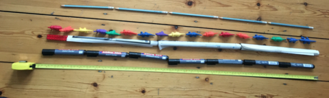 How long are you? Various items arranged in a line on a wooden floor. From top to bottom, there are five pencils lying end-to-end, 17 coloured toy dinosaurs, three rolls of paper and a lego brick, seven whiteboard pens, and a tape measure.