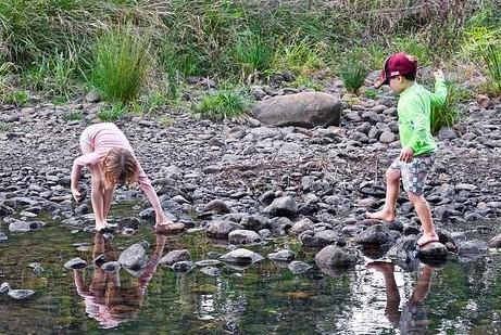 Paths Two children playing by a shallow rocky stream. One child is bending down to pick up stones, while the other steps barefoot on rocks. Both are surrounded by stones and greenery in the background.