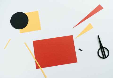 Making a Picture A selection of colourful paper cut-outs on a white surface, including a red rectangle, black circle, yellow square, and various triangular and strip shapes. A pair of black scissors is placed on the right side.