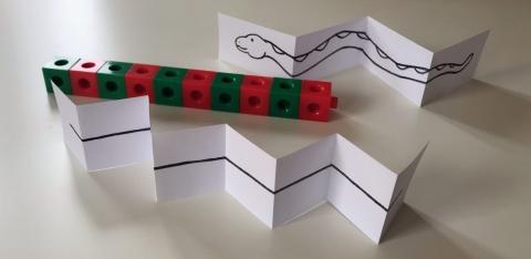 Long Creatures A line of multilink cubes on a table, alternating between red and green. On either side are two pieces of card, folded to look like snakes.