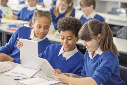 Three primary school students wearing blue sitting at a table all looking at iPads