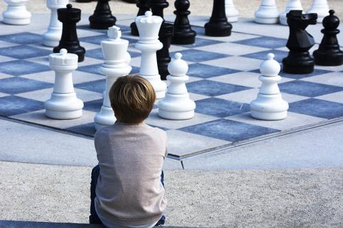 Outdoor chess in the background, boy sitting in foreground with back to camera, looking at the chess pieces