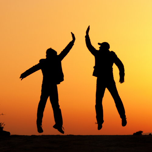 The silhouettes of two people jumping and high fiving at sunset.