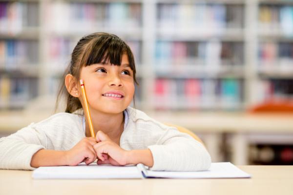 Young girl smiling while looking up and to her left, holding a pencil and sitting at a table with a notebook