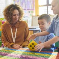 A teacher at a table with young children, looking at a colourful grid and dice.